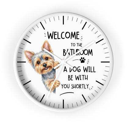 A round white wall clock with a black hand-drawn style outline frame, black hour markers, and a printed illustration of a Yorkshire Terrier peeking from behind the word 'BATHROOM' in a humorous quote that reads 'WELCOME TO THE BATHROOM A DOG WILL BE WITH YOU SHORTLY'.