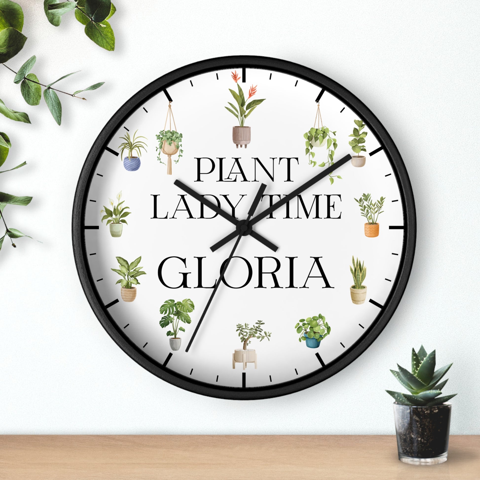 A round wall clock with a black frame and a white face, featuring the text 'PLANT LADY TIME GLORIA' and illustrated potted plants around the dial. The clock is mounted on a white wall above a light wood surface, with decorative green leaves and a small succulent nearby.