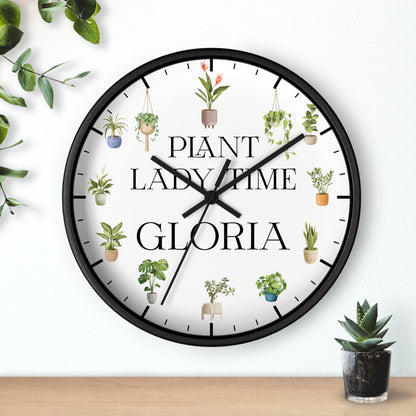 A round wall clock with a black frame and a white face, featuring the text 'PLANT LADY TIME GLORIA' and illustrated potted plants around the dial. The clock is mounted on a white wall above a light wood surface, with decorative green leaves and a small succulent nearby.