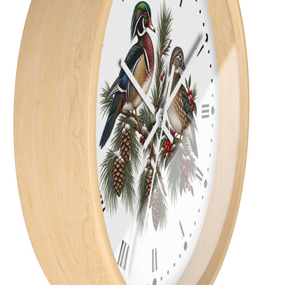 A close-up of a round wall clock with a light natural wood frame and a white face featuring an illustrated woodland scene with two birds perched on pine branches with red berries.