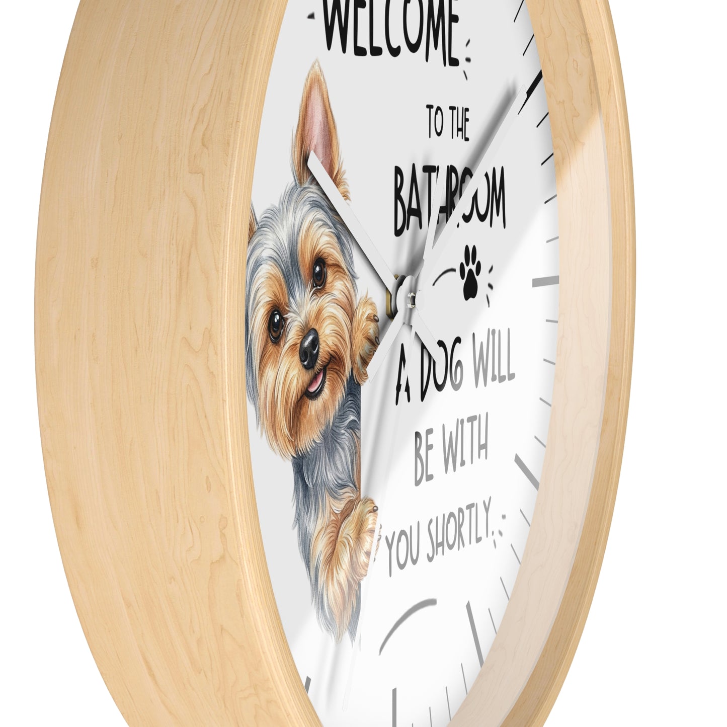 A round wall clock with a light natural wood frame and a white face. The clock features a hand-drawn illustration of a Yorkshire Terrier peeking from behind text that reads 'WELCOME TO THE BATHROOM A DOG WILL BE WITH YOU SHORTLY'.