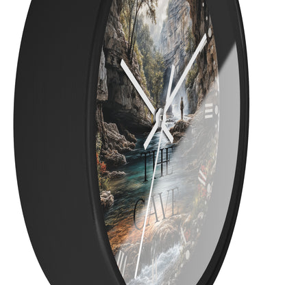 A round wall clock with a black frame and a scenic landscape print showing a cave interior, river, trees, and the words “THE CAVE,” photographed against a white background.