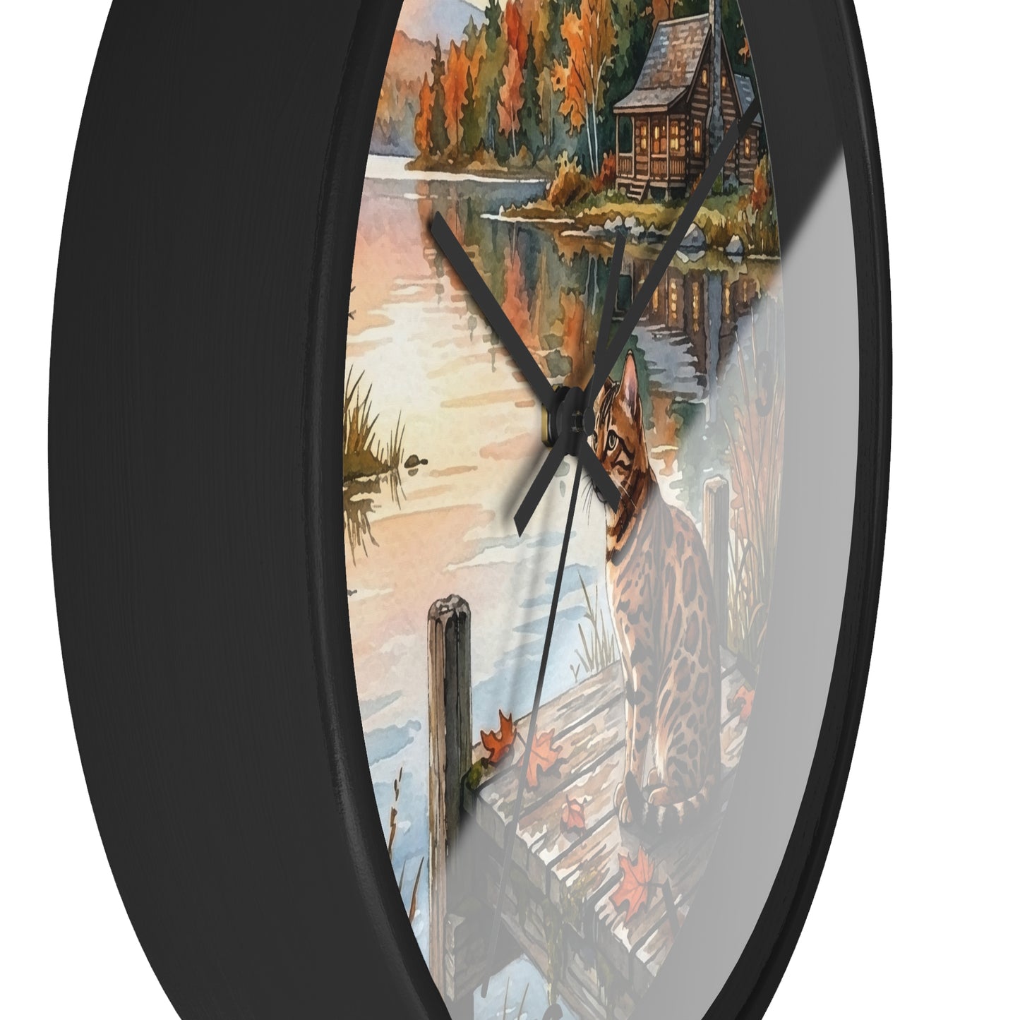 A round wall clock with a black frame and a scenic printed face showing a lake, autumn trees, a cabin, and a dock. The clock is photographed against a plain white background.