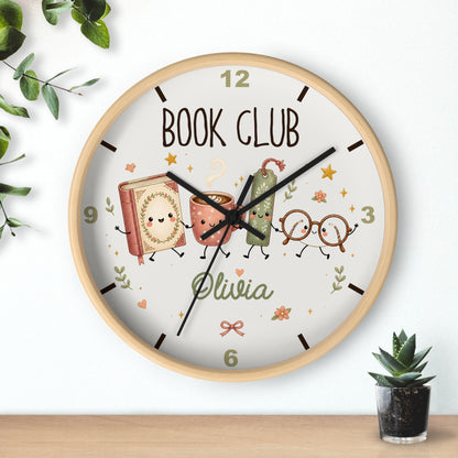 A round wall clock with a light wood frame and a whimsical 'Book Club' design on the dial. The clock face features cartoon illustrations of a book, a coffee cup, a pencil, and reading glasses, along with the name 'Olivia' in green script.