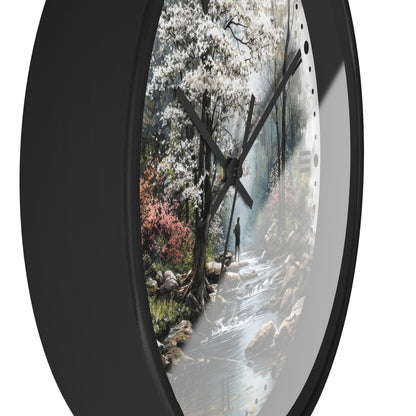Round wall clock with a black frame and a printed scenic dial showing a forest stream, white blossoms, and a small figure, photographed against a white background.