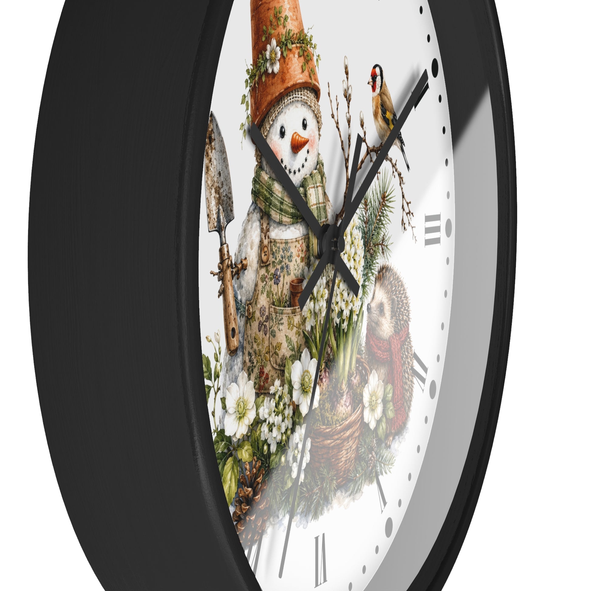 A round wall clock with a black frame and a white face featuring a winter garden illustration. The design shows a snowman wearing a terracotta pot hat and scarf, holding a trowel, with a small bird perched on a branch above.