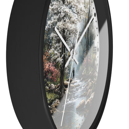 Round wall clock with a black frame and a printed scenic face showing a forest stream, white blossoms, and a small figure, photographed against a white background.