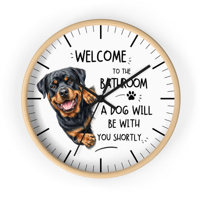 A round wall clock with a white face, black hands, black hour markers, and a printed illustration of a Rottweiler dog next to the humorous text, 'WELCOME TO THE BATHROOM A DOG WILL BE WITH YOU SHORTLY.'.