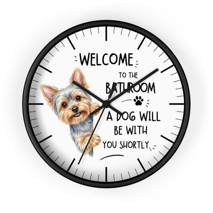A round wall clock with a black frame and white face, featuring a small dog illustration and the text 'WELCOME TO THE BATHROOM A DOG WILL BE WITH YOU SHORTLY.' on a light gray background.