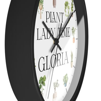 A close-up of a round wall clock with a black frame and a white face featuring the text 'PLANT LADY TIME GLORIA' and small potted plant illustrations.