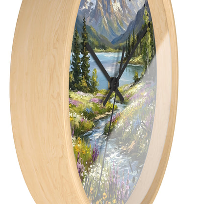 Round wall clock with a light natural wood frame and a scenic landscape print featuring a mountain lake, pine trees, and a winding stream with wildflowers.