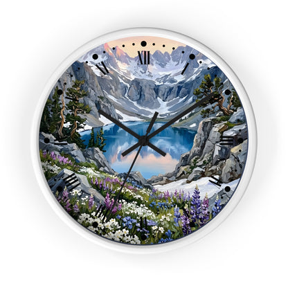 A round wall clock with a white frame and a scenic mountain landscape printed on the face. The artwork shows snow-capped peaks, a blue alpine lake, rocky cliffs, evergreen trees, and wildflowers, with black Roman numerals and black clock hands.
