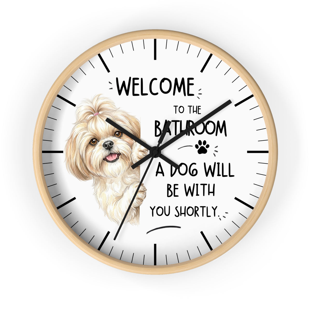 A round wall clock with a light wood frame and a white face, featuring a small fluffy cream-colored dog illustration and the text 'WELCOME TO THE BATHROOM A DOG WILL BE WITH YOU SHORTLY.' The clock is shown against a plain white background.