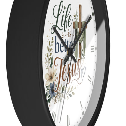 A close-up of a round wall clock with a black frame and a white face featuring the phrase 'Life is better with Jesus' in decorative script, along with floral and cross illustrations.