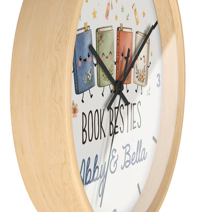 A round wall clock with a light natural wood frame and a white face featuring four smiling illustrated books and the text 'BOOK BESTIES Abby & Bella'.