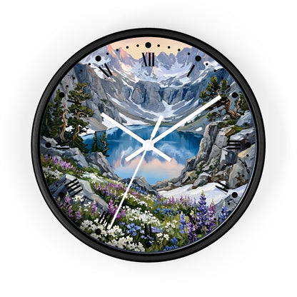 A round wall clock with a black frame and a scenic mountain landscape print on the face, featuring a blue lake, snowy peaks, rocky cliffs, and wildflowers. The clock has black Roman numeral hour markers and white hands, shown against a plain white background.