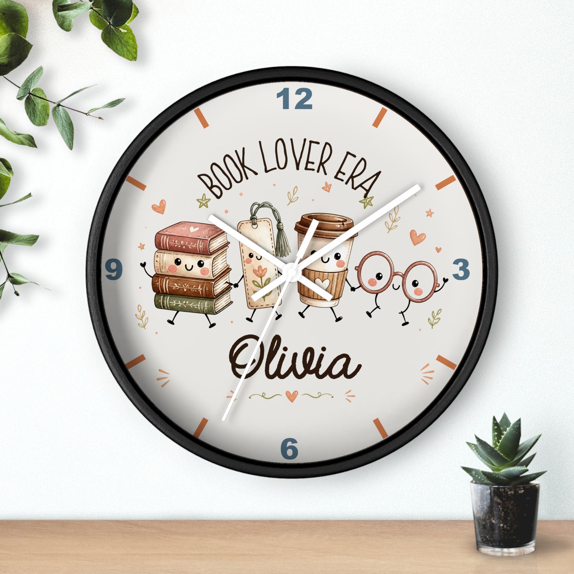 A round wall clock with a black frame and a white face, decorated with cute book and coffee cup characters and the text 'BOOK LOVER ERA' and the name 'Olivia'. The clock is shown hanging on a white wall above a wooden surface, with small green plants in the corners.