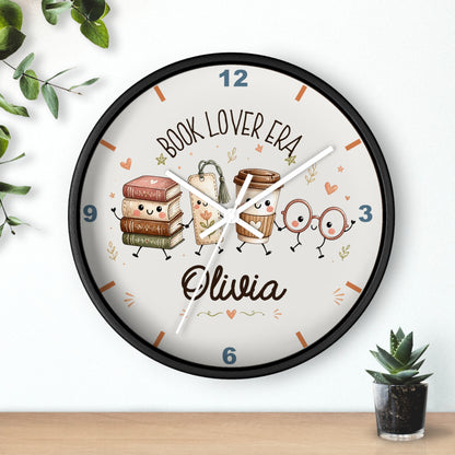 A round wall clock with a black frame and a white face, decorated with cute book and coffee cup characters and the text 'BOOK LOVER ERA' and the name 'Olivia'. The clock is shown hanging on a white wall above a wooden surface, with small green plants in the corners.
