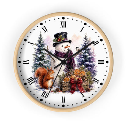 A round wall clock with a light wood frame and a white face featuring a winter holiday illustration of a snowman, a squirrel, evergreen trees, and a gift basket. The clock has black Roman numerals and hands on a clean white background.