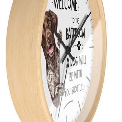 A round wall clock with a light natural wood frame and a white face. The clock features a German shorthaired pointer dog illustration on the left and black text that reads 'WELCOME TO THE BATROOM A DOG WILL BE WITH YOU SHORTLY'.
