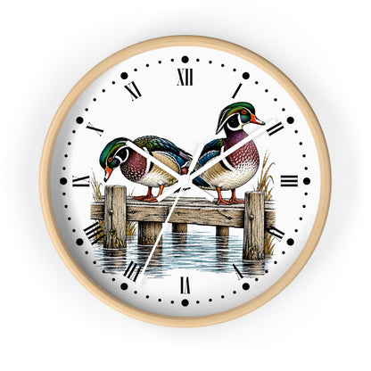 A round wall clock with a light natural wood frame and a white face featuring an illustration of two mallards on a wooden dock over water, with black Roman numerals and hands.
