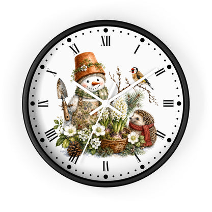 A round wall clock with a black frame and a white face featuring a whimsical winter garden illustration. The design includes a snowman in a terracotta pot hat, a small bird, a hedgehog in a red scarf, white flowers, pinecones, and gardening tools, with black Roman numerals and white hands.
