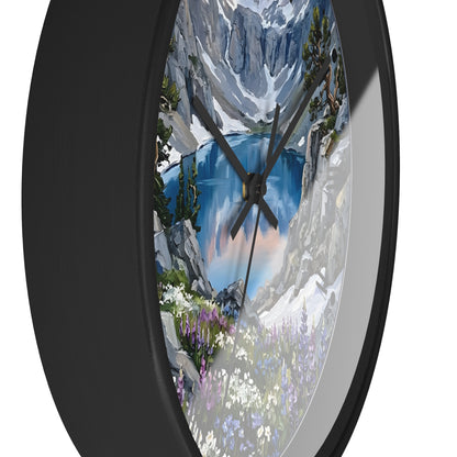A round wall clock with a black frame and a scenic landscape print featuring a blue lake, snowy mountains, trees, and wildflowers, shown against a plain white background.