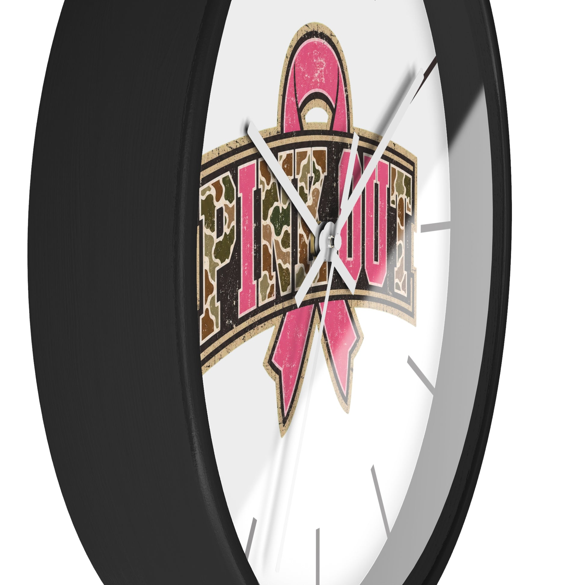 A close-up of a round wall clock with a black frame and a white face, featuring a large pink ribbon and camouflage-style text design on the front.