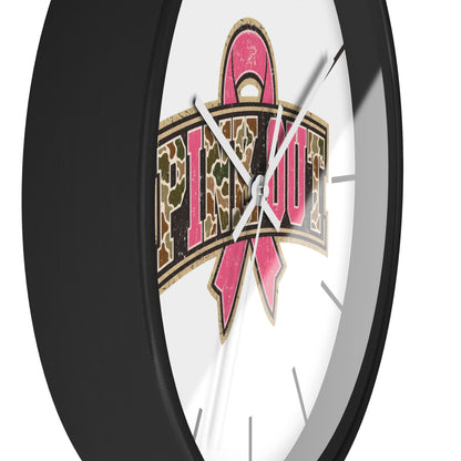 A close-up of a round wall clock with a black frame and a white face, featuring a large pink ribbon and camouflage-style text design on the front.
