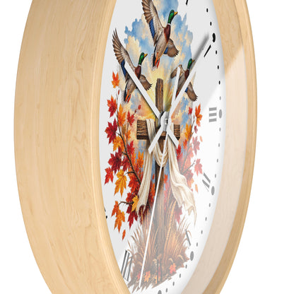 Round wall clock with a light natural wood frame and a white face featuring a hunting scene with mallards, autumn leaves, a cross, and a draped white cloth.