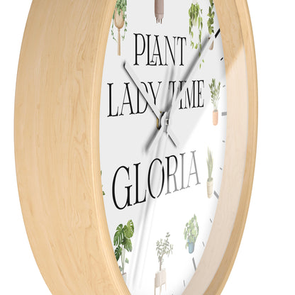 Round wall clock with a light natural wood frame and a white face featuring the text 'PLANT LADY TIME GLORIA' and small potted plant illustrations.