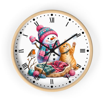 A round wall clock with a light wood frame and a white face, decorated with a festive winter illustration featuring a snowman in a pink and blue knit hat, an orange tabby cat, a small bird, yarn balls, and a plaid blanket.