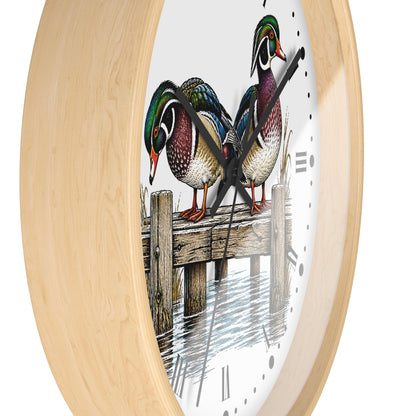 Round wall clock with a light natural wood frame and a white face featuring an illustration of three wood ducks standing on a wooden dock over water, with black Roman numerals and black hands.