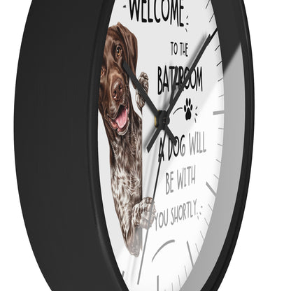 A round wall clock with a black frame and a white face featuring a German shorthaired dog illustration and the text 'WELCOME TO THE BATHROOM A DOG WILL BE WITH YOU SHORTLY'.