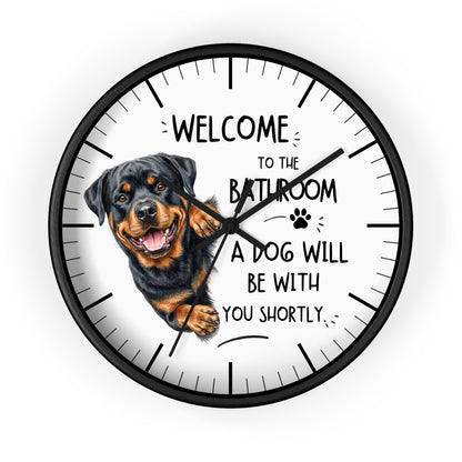 A round wall clock with a black frame and a white face featuring a Rottweiler illustration and the text 'WELCOME TO THE BATHROOM A DOG WILL BE WITH YOU SHORTLY.' on a light gray background.