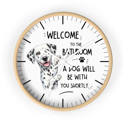 A round wall clock with a light wood frame and a white face, featuring a watercolor illustration of a Dalmatian dog and the text 'WELCOME TO THE BATHROOM A DOG WILL BE WITH YOU SHORTLY.' on a plain white background.