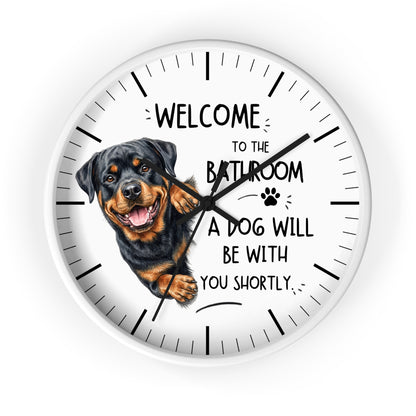 A round wall clock with a white face, black hands, black hour markers, and a printed illustration of a Rottweiler dog next to the humorous text, 'WELCOME TO THE BATHROOM A DOG WILL BE WITH YOU SHORTLY.'.