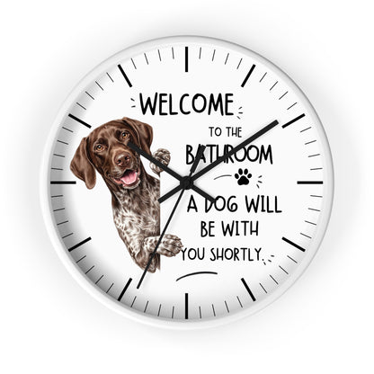 A round white wall clock with black hands and hour markers, featuring a brown and white pointer dog illustration and the text 'WELCOME TO THE BATHROOM A DOG WILL BE WITH YOU SHORTLY.' on a white background.