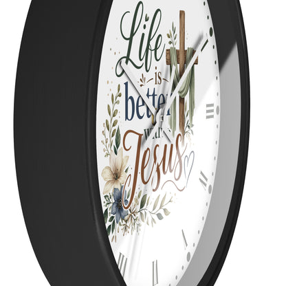 A close-up of a round wall clock with a black frame and a white face featuring the phrase 'Life is better with Jesus' in decorative script, along with floral illustrations and a wooden cross.