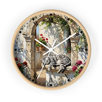 Round wall clock with a light wood frame and a printed scenic dial showing a gray tabby cat resting on a stone ledge beneath an arched window with flowers and shadows.