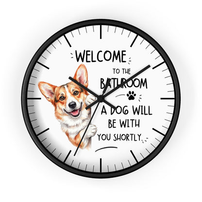 A round wall clock with a black frame and white face, featuring a cartoon corgi illustration and the text 'WELCOME TO THE BATHROOM A DOG WILL BE WITH YOU SHORTLY.' on a light gray background.