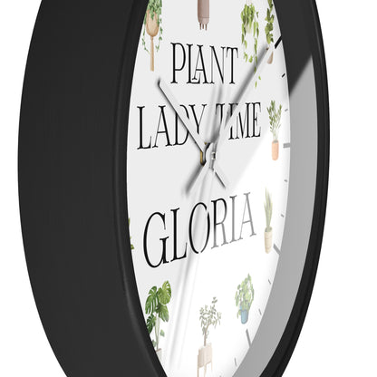 A close-up of a round wall clock with a black frame and a white face featuring the text 'PLANT LADY TIME GLORIA' and small cactus and plant illustrations.