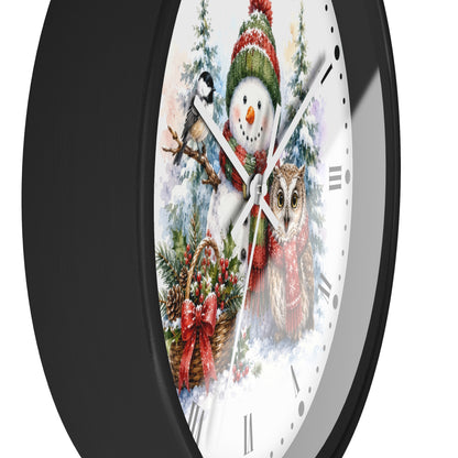 A round wall clock with a black frame and a white face featuring a festive winter illustration of a snowman, bird, owl, pine trees, and a basket with red bows.