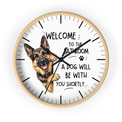 A round wall clock with a light wood frame and a white face, featuring a German Shepherd illustration and the text 'WELCOME TO THE BATHROOM A DOG WILL BE WITH YOU SHORTLY.' on a plain white background.