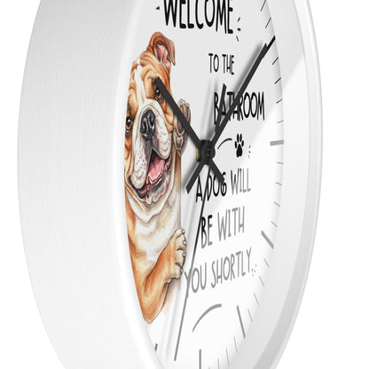 A white round wall clock with a humorous dog-themed design. The clock face features a detailed illustration of a brown and white bulldog peeking from behind text that reads 'WELCOME TO THE BATHROOM A DOG WILL BE WITH YOU SHORTLY.