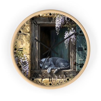 Round wall clock with a light wood frame and a printed illustration of a gray cat sleeping on a rustic wooden windowsill, surrounded by wisteria flowers and stone-textured details. The clock face includes black Roman numerals and black clock hands.