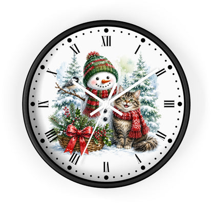 A round wall clock with a black frame and a festive winter illustration on the face. The design shows a snowman in a green and red knit hat and scarf beside a gray tabby cat wearing a red scarf, with snowy trees, a basket of greenery, and red bows in the background.