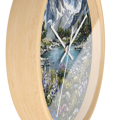 An oval wall clock with a light natural wood frame and a scenic landscape print featuring a mountain lake, wildflowers, and trees.