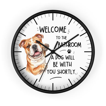 A round wall clock with a black frame and a white face, featuring a brown and white bulldog illustration and the text 'WELCOME TO THE BATHROOM A DOG WILL BE WITH YOU SHORTLY.' on a light gray background.