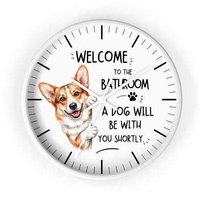 A round white wall clock with black hands and black hour markers, featuring a printed illustration of a corgi dog and the text 'WELCOME TO THE BATHROOM A DOG WILL BE WITH YOU SHORTLY.' on a white background.